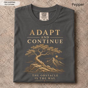 K&ouml;nnte beinhalten: Ein anthrazitfarbenes T-Shirt mit einer goldenen Grafik eines Bonsai-Baums auf einem Felsen und Bergen im Hintergrund. Der Text lautet ADAPT AND CONTINUE und THE OBSTACLE IS THE WAY. Das T-Shirt hat ein Etikett der Marke Comfort Colors.