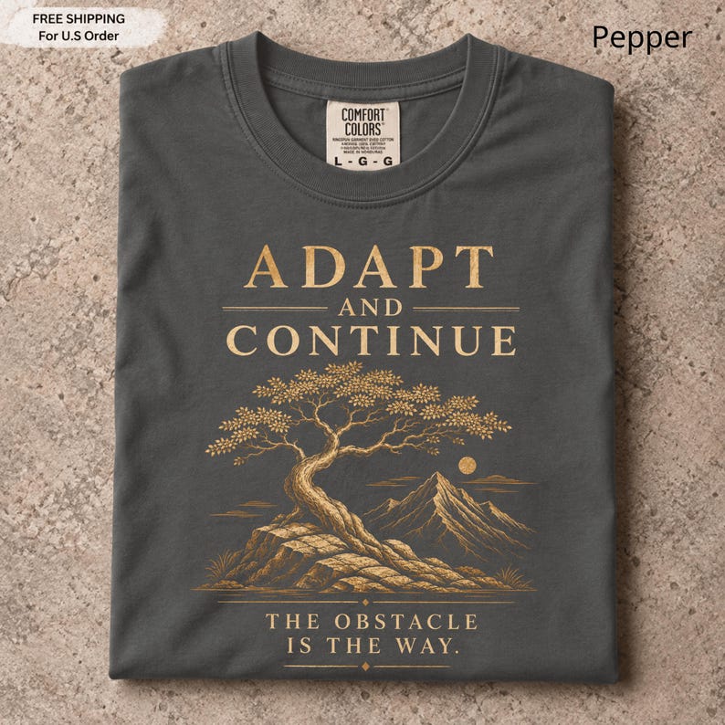 K&ouml;nnte beinhalten: Ein anthrazitfarbenes T-Shirt mit einer goldenen Grafik eines Bonsai-Baums auf einem Felsen und Bergen im Hintergrund. Der Text lautet ADAPT AND CONTINUE und THE OBSTACLE IS THE WAY. Das T-Shirt hat ein Etikett der Marke Comfort Colors.