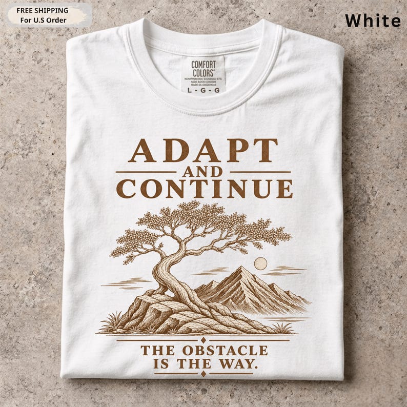 K&ouml;nnte beinhalten: Ein wei&szlig;es T-Shirt mit einem braunen Grafikdruck eines Bonsai-Baums auf einem Felsen mit Bergen im Hintergrund. Der Text ADAPT AND CONTINUE steht &uuml;ber dem Baum und THE OBSTACLE IS THE WAY unter dem Baum.