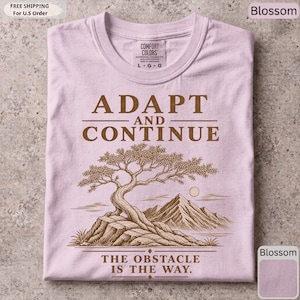 K&ouml;nnte beinhalten: Ein hellrosa T-Shirt mit einem braunen Grafikdruck eines Bonsai-Baums auf einem Felsvorsprung mit Bergen im Hintergrund. Der Text lautet ADAPT AND CONTINUE &uuml;ber dem Baum und THE OBSTACLE IS THE WAY unter dem Baum.