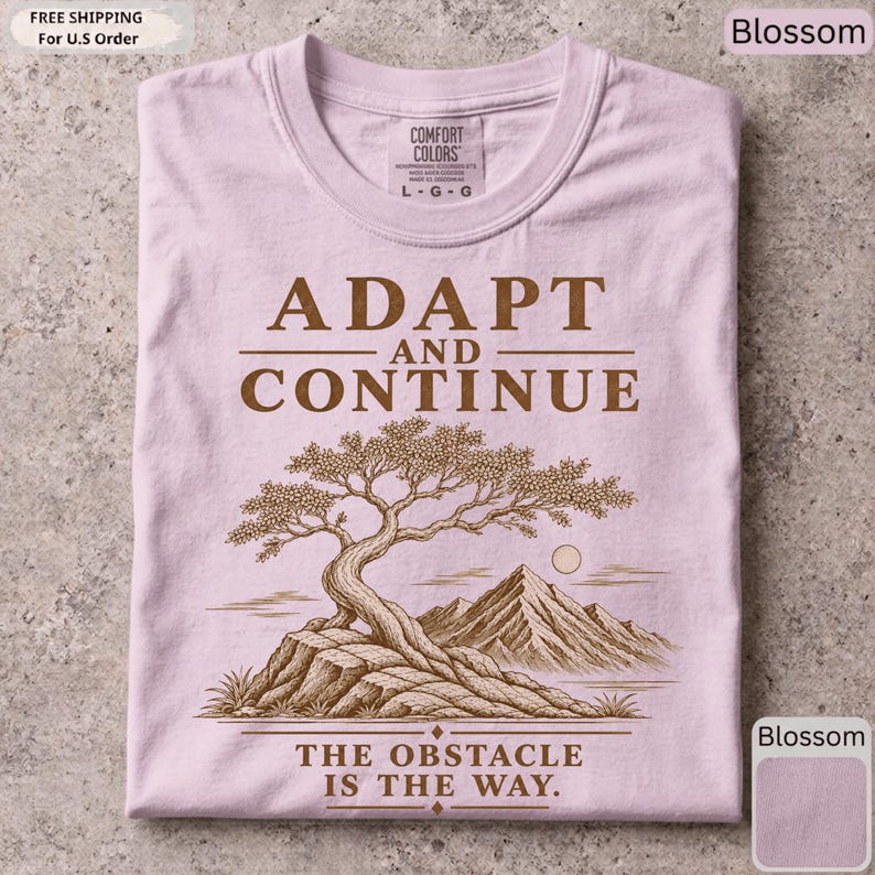 K&ouml;nnte beinhalten: Ein hellrosa T-Shirt mit einem braunen Grafikdruck eines Bonsai-Baums auf einem Felsvorsprung mit Bergen im Hintergrund. Der Text lautet ADAPT AND CONTINUE &uuml;ber dem Baum und THE OBSTACLE IS THE WAY unter dem Baum.