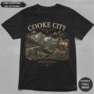 May include: A black t-shirt featuring a graphic print of a mountain landscape with a bighorn sheep, a winding road, and text that reads COOKE CITY MONTANA, BEARTOOTH HIGHWAY ELEV 10,947 FT, and GATEWAY TO BEARTOOTH PASS ELEV 10,947 FT.