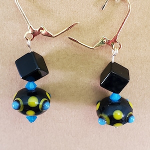 Può includere: Un paio di orecchini fatti a mano con perline a cubo nere e perline rotonde nere con dettagli gialli e blu. Gli orecchini hanno ganci per le orecchie dorati, che mostrano un design unico e artistico.