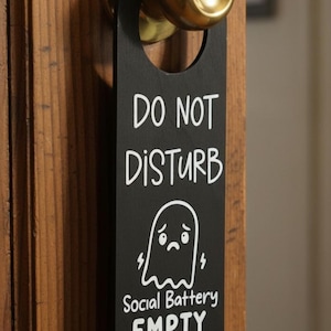 May include: Black door hanger with the text "DO NOT DISTURB" and "Social Battery EMPTY" in white. A sad ghost illustration is also featured. The hanger is hanging on a wooden door with a gold doorknob.
