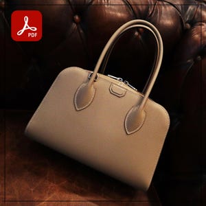 May include: A tan leather handbag with a rectangular shape and rounded corners. The bag features two top handles and a silver-toned clasp. White stitching accents the edges and decorative elements. The bag is set against a brown leather background.