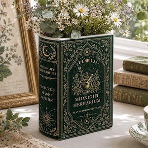 May include: A dark green ceramic vase shaped like a vintage book titled Midnight Herbarium. The front features gold foil illustrations of a mortar and pestle, moon phases, and stars. It is filled with a bouquet of white daisies and eucalyptus.
