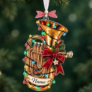 Op de afbeelding: Een goudkleurige tuba kerstornament versierd met kleurrijke kerstverlichting, een rode strik, hulstbessen en een dennenappel. Een wit lint met een rood-wit geblokte strik hangt aan de bovenkant. Een banner onderaan bevat de tekst Name.