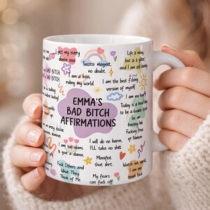 May include: White ceramic mug with the text "EMMA'S BAD BITCH AFFIRMATIONS" in a purple bubble. The mug is covered in affirmations and doodles in various colors. The mug is held by a person wearing a cream-colored sweater.