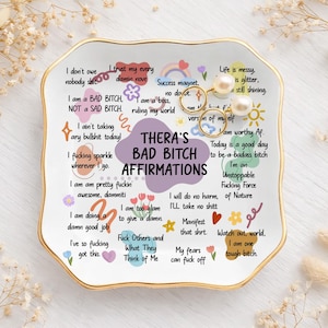 Könnte beinhalten: Eine weiße Keramik-Schmuckschale mit goldenem Rand, mit dem Text "Thera's Bad Bitch Affirmations" und verschiedenen Affirmationen im handschriftlichen Stil. Die Schale enthält zwei Ringe und ein Paar Perlenohrringe.