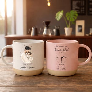 Op de afbeelding: Twee keramische mokken met een beige basis en een witte handgreep. Eén mok heeft een tekening van een vader en kind met de tekst "Daddy & Aurora". De andere mok is roze met een klokontwerp en de tekst "The moment you became Dad."