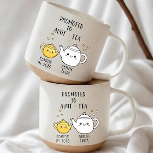Könnte beinhalten: Zwei weiße Keramikbecher mit dem Text "PROMOTED TO AUNT - TEA" und den Worten "COMING IN 2026" und "AUNTIE LUNA". Jeder Becher zeigt ein Cartoon-Teekannen- und Tassendesign mit Herzakzenten. Die Becher haben einen beigefarbenen Boden.