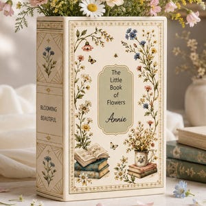 May include: A rectangular ceramic vase shaped like a book, featuring floral illustrations, butterflies, and the text "The Little Book of Flowers Annie" and "BLOOMING BEAUTIFUL." The vase is filled with a bouquet of blue, pink, and white flowers.