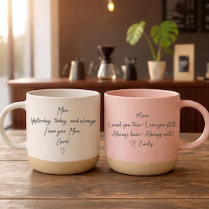 Puede incluir: Dos tazas de cerámica con acabado crema y rosa, cada una con base beige y asa curva. La taza crema tiene el texto "Mom Yesterday, today, and always I love you, Mom Carol ♡". La taza rosa tiene el texto "Mom Loved you then - Love you still Always have - Always will! ♡ Emily".