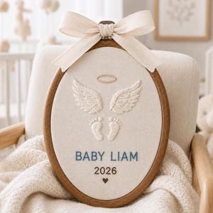 Peut inclure: Un tambour à broder ovale en bois contenant un tissu crème avec des ailes d'ange blanches brodées, une auréole et des empreintes de pieds de bébé. Le texte indique BABY LIAM et 2026 avec un petit cœur. Un ruban beige est noué en haut.