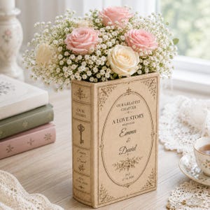 May include: A rectangular beige book-shaped vase featuring the text Our Greatest Chapter A Love Story Written By Emma & David Est 07.01.2026, filled with a bouquet of light pink roses, white roses, and small white baby's breath flowers.
