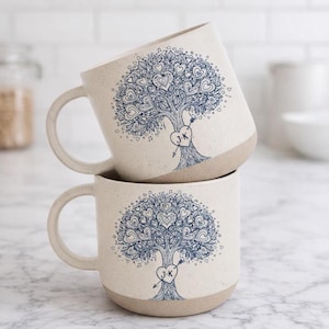 Può includere: Due tazze in ceramica impilate con finitura screziata color crema. Ogni tazza presenta un disegno di albero blu pieno di foglie a forma di cuore e un cuore con iniziali. La base di ogni tazza è di colore marrone chiaro.