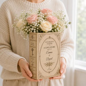 May include: A rectangular book-shaped vase in a light beige finish, featuring decorative black text and filigree borders. The front reads 'OUR GREATEST CHAPTER A LOVE STORY WRITTEN BY Emma & David EST. 07.01.2026'. It holds a bouquet of pink roses and baby's breath.