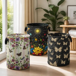 May include: Three cylindrical fabric storage baskets with black side handles. The left basket features a floral and butterfly print on a striped background. The center basket displays a celestial sun and flower motif. The right basket has a quilted butterfly pattern.