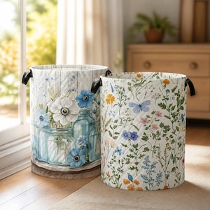 May include: Two cylindrical fabric laundry hampers with quilted textures and black side handles. One features a blue floral print with glass jars on a wood background, while the other displays a colorful wildflower and butterfly pattern on white.