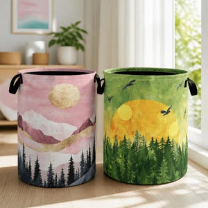 May include: Two cylindrical fabric laundry hampers with black side handles. The left hamper features a pink mountain landscape with a gold sun and dark pine trees. The right hamper displays a green forest scene with a yellow sun and flying birds.