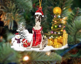 Peut inclure: Ornement de Noël représentant un Grand Danois portant un bonnet de Père Noël et une écharpe rouge. Le chien est entouré d'un bonhomme de neige, de cadeaux et d'un sapin de Noël décoré. L'ornement est suspendu à une ficelle dorée.