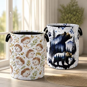 May include: Two cylindrical fabric laundry hampers with black handles. The left hamper features a repeating pattern of brown hedgehogs and green leaves. The right hamper displays a large bear silhouette filled with a forest and mountain scene.