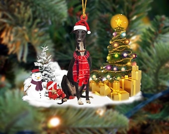 May include: A Christmas ornament featuring a black dog wearing a Santa hat and a red scarf. The dog is surrounded by a Christmas tree, presents, and a snowman. The ornament hangs from a gold string.