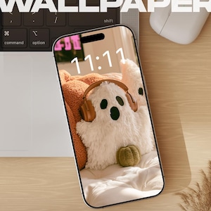Puede incluir: Un smartphone que muestra un fondo de pantalla digital de un peluche blanco y peludo con forma de fantasma que lleva auriculares marrones, con la hora 11:11 visible. El teléfono está sobre un escritorio junto a un teclado, un ratón blanco y hierba seca.