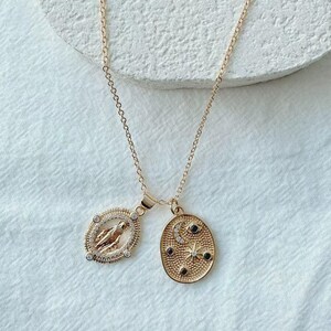 May include: Gold-toned necklace featuring two pendants. One pendant is an oval with a religious figure, surrounded by clear stones. The other pendant is an oval with a crescent moon, stars, and black stones. The necklace hangs on a delicate chain.