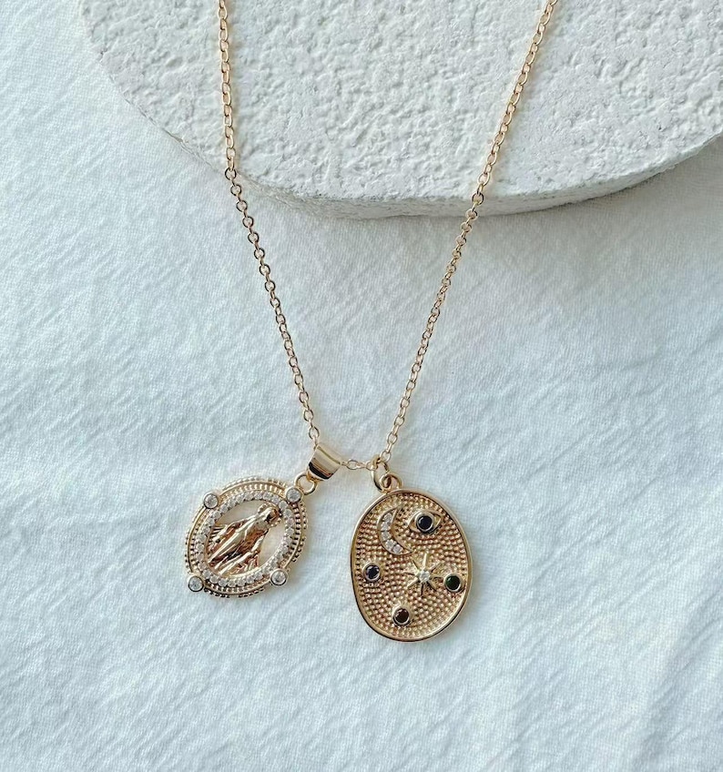 May include: Gold-toned necklace featuring two pendants. One pendant is an oval with a religious figure, surrounded by clear stones. The other pendant is an oval with a crescent moon, stars, and black stones. The necklace hangs on a delicate chain.