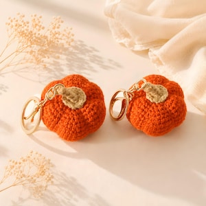 May include: Two crochet pumpkin keychains featuring vibrant orange yarn bodies and light tan stems. Each pumpkin is attached to a polished gold-tone metal split ring by a matching gold-tone chain, showcasing a textured, hand-stitched appearance.
