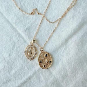 May include: Two gold-colored necklaces with pendant charms. One pendant is an oval shape with a Virgin Mary design, and the other is an oval shape with a crescent moon, stars, and small black and clear stones. The necklaces are on a white fabric background.
