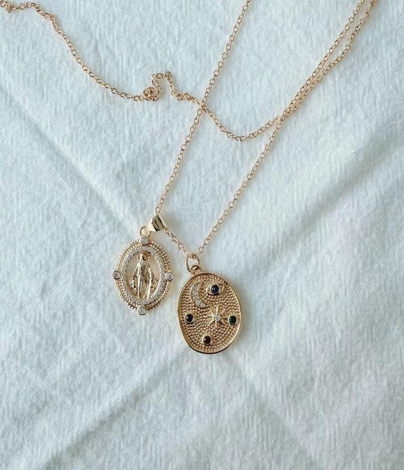 May include: Two gold-colored necklaces with pendant charms. One pendant is an oval shape with a Virgin Mary design, and the other is an oval shape with a crescent moon, stars, and small black and clear stones. The necklaces are on a white fabric background.