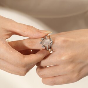 May include: A silver ring featuring a large, oval, pale pink gemstone. The ring has an openwork design with smaller, clear gemstones. The ring is being held by a person.
