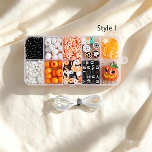 Può includere: Una scatola organizer in plastica trasparente etichettata Style 1 contenente perline assortite a tema Halloween, tra cui perline rotonde nere, bianche e arancioni, dischi in argilla, cubi con lettere, stelle e un ciondolo a forma di zucca, con un cordino bianco.