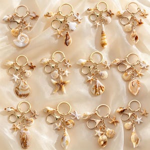 May include: A collection of gold-colored keychains, each adorned with a variety of seashells and starfish charms. The keychains feature different shell shapes and sizes, with a mix of natural colors like beige, brown, and white. The charms are attached with small gold rings.