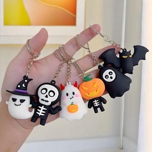May include: A set of five Halloween-themed keychains featuring silicone charms: a ghost in a witch hat, a skeleton, a ghost with red horns holding a pumpkin, a pumpkin-headed figure, and a black bat with a ghost, all attached to metal rings.