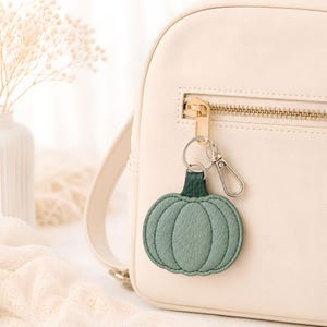 Peut inclure: Un porte-clés en forme de citrouille vert sauge avec une tige vert foncé, fixé à un mousqueton et un anneau en métal argenté. Le porte-clés est accroché à la tirette de fermeture éclair dorée d'un sac à dos de couleur crème à la finition lisse.