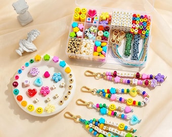 May include: A clear plastic organizer box filled with assorted colorful beads, including letter cubes, smiley faces, hearts, and flowers. Beside it, a white circular tray holds loose beads, and four finished beaded keychains with gold clasps.