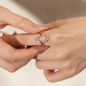 May include: Rose gold ring featuring a central oval pink gemstone, encircled by smaller clear stones and baguette-cut accents. The ring is set on a delicate band, displaying a cluster design. The ring is worn on a finger.