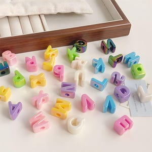 May include: A collection of small, multi-coloured alphabet beads scattered on a surface. The beads feature a glossy finish in various shades including pink, yellow, blue, purple, green, white, and iridescent metallic tones, each with a central hole.