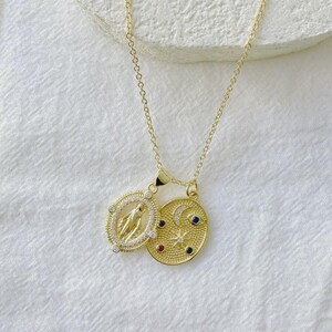 May include: Gold-toned necklace featuring two pendants. One pendant is oval-shaped with a religious figure, surrounded by small clear stones. The other pendant is an oval with a crescent moon, stars, and colorful gemstones.