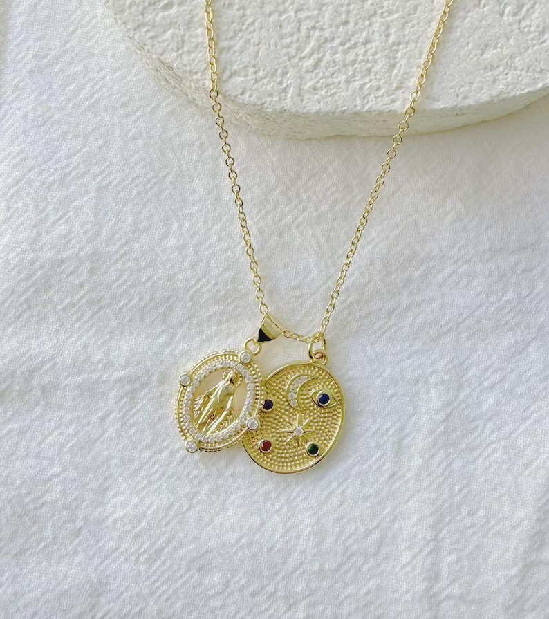 May include: Gold-toned necklace featuring two pendants. One pendant is oval-shaped with a religious figure, surrounded by small clear stones. The other pendant is an oval with a crescent moon, stars, and colorful gemstones.