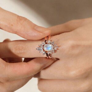 May include: Rose gold ring featuring an oval moonstone at its centre, encircled by a halo of small, clear, baguette-cut and round-cut gemstones. The ring sits on a slender band, creating a sparkling, elegant design. The ring is shown on a person's finger.