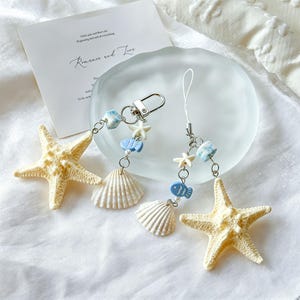 Puede incluir: Dos llaveros con temática de playa con estrellas de mar, conchas marinas y dijes de peces azules. Uno tiene un cierre plateado, el otro un lazo blanco. Una estrella de mar y una tarjeta con el texto "Romance and Love" están en el fondo.