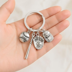 May include: A silver-toned metal keyring featuring four baseball-themed charms attached to a circular split ring. The charms include a round baseball with stitched detailing, a slender baseball bat, a baseball glove holding a ball, and a baseball cap.