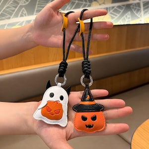 Könnte beinhalten: Zwei Halloween-Schlüsselanhänger aus Leder, bestehend aus einem weißen Geist mit einem orangefarbenen Kürbis und einem orangefarbenen Kürbis mit schwarzem Hexenhut. Beide Anhänger sind an schwarzen geflochtenen Kordeln mit gelben Akzenten befestigt.