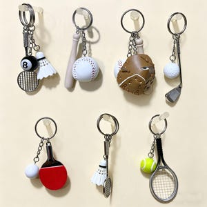 May include: A collection of seven metal keychains featuring miniature sports equipment, including a tennis racket with a shuttlecock and an 8-ball, a baseball bat with a ball, a baseball glove, a golf club with a ball, a table tennis paddle, and a tennis racket with a ball.