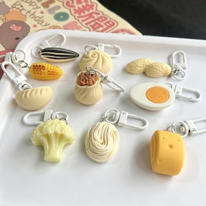 May include: A collection of food-themed keychains, including a sunflower seed, corn on the cob, dumpling, bao bun, peanut, egg, cauliflower, noodles, and cheese. Each keychain has a silver clasp.