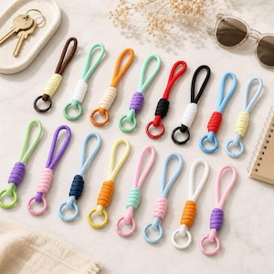 May include: A collection of sixteen colorful cord keychains, each featuring a looped cord, a wrapped knot section in a contrasting color, and a small circular ring at the base. The keychains are arranged in two rows and display various color combinations.
